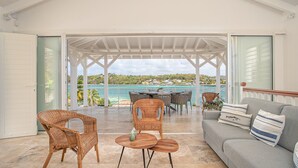 A covered outdoor seating area with wicker furniture, a grey sofa with striped pillows, and a view of the sea and distant houses. - Blue Cap (Le François)