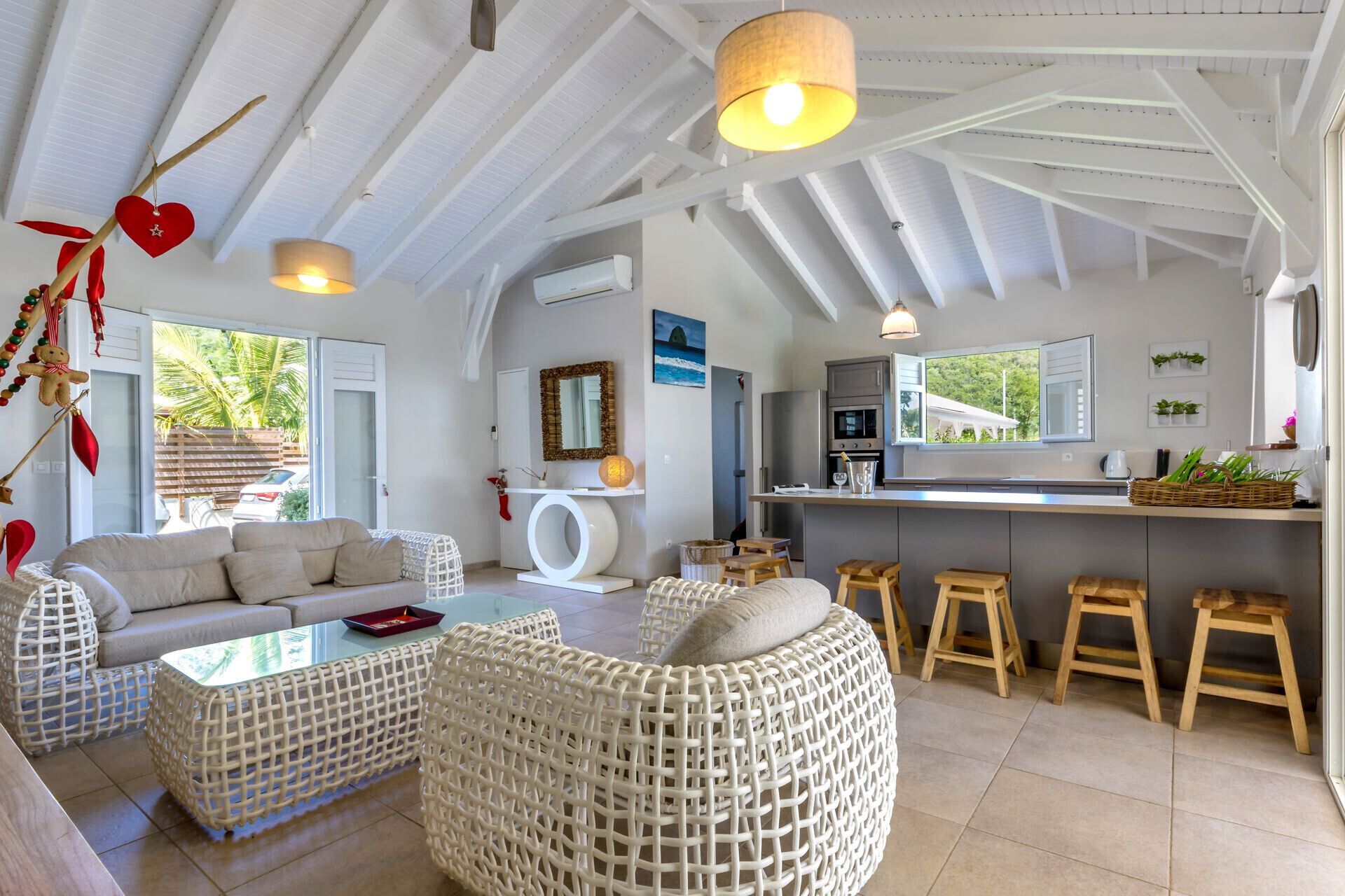 A modern living room with a sofa, wicker ottoman, and a glass coffee table. There is a kitchen area with a counter and stools.