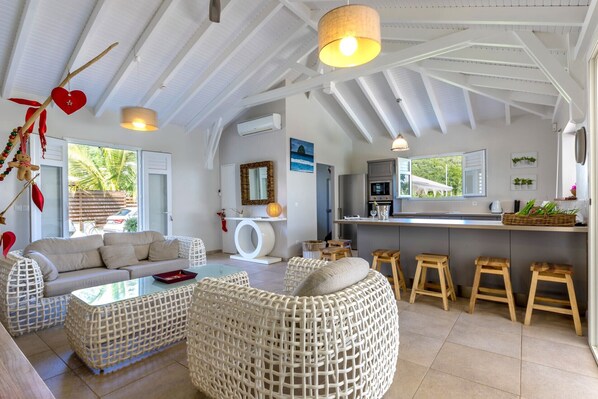 A modern living room with a sofa, wicker ottoman, and a glass coffee table. There is a kitchen area with a counter and stools.