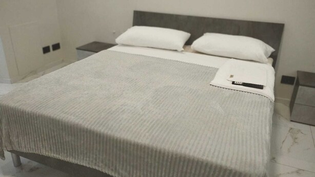 A neatly made bed with a grey bedspread and two white pillows, a bedside table with a lamp, and a small nightstand. - Bnb Massaua (Torino)