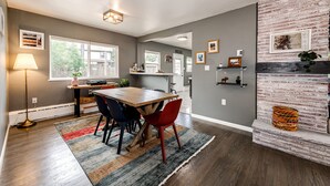 A modern kitchen with a wooden table, chairs, and a red accent chair. There is a large window providing natural light, and the walls are painted gray. - Relax & Recharge – Spacious Wheatridge Stay w/ Backyard Fun (Wheat Ridge)