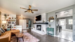 A modern living room with a fireplace, a ceiling fan, and a kitchen area visible through an open door. - Relax & Recharge – Spacious Wheatridge Stay w/ Backyard Fun (Wheat Ridge)