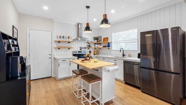 A modern kitchen with white cabinetry, a stainless steel refrigerator, wooden bar stools, pendant lighting, and a window providing natural light.