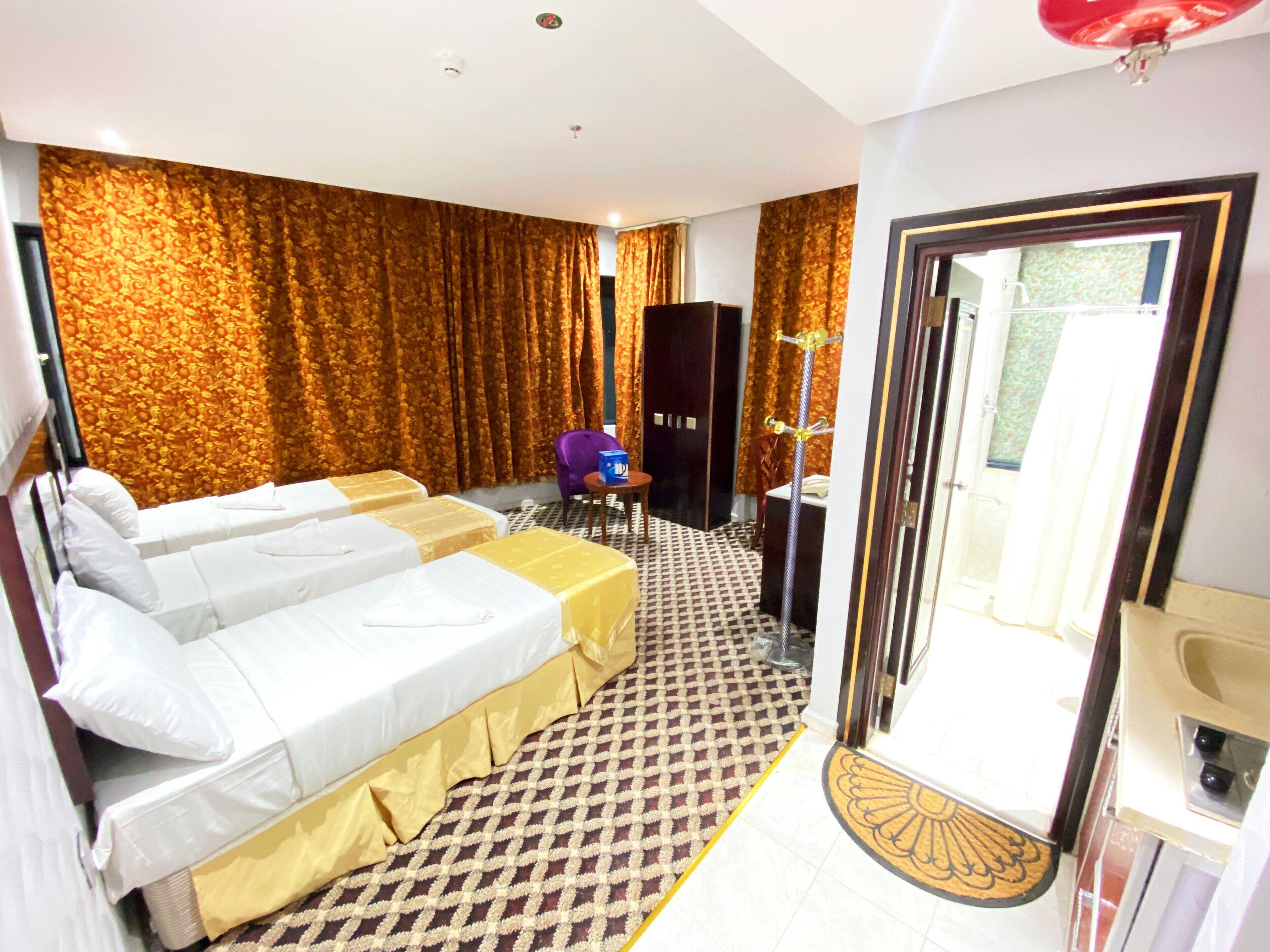 A hotel room with two beds, a desk, a chair, a wardrobe, and a bathroom with a sink and mirror.