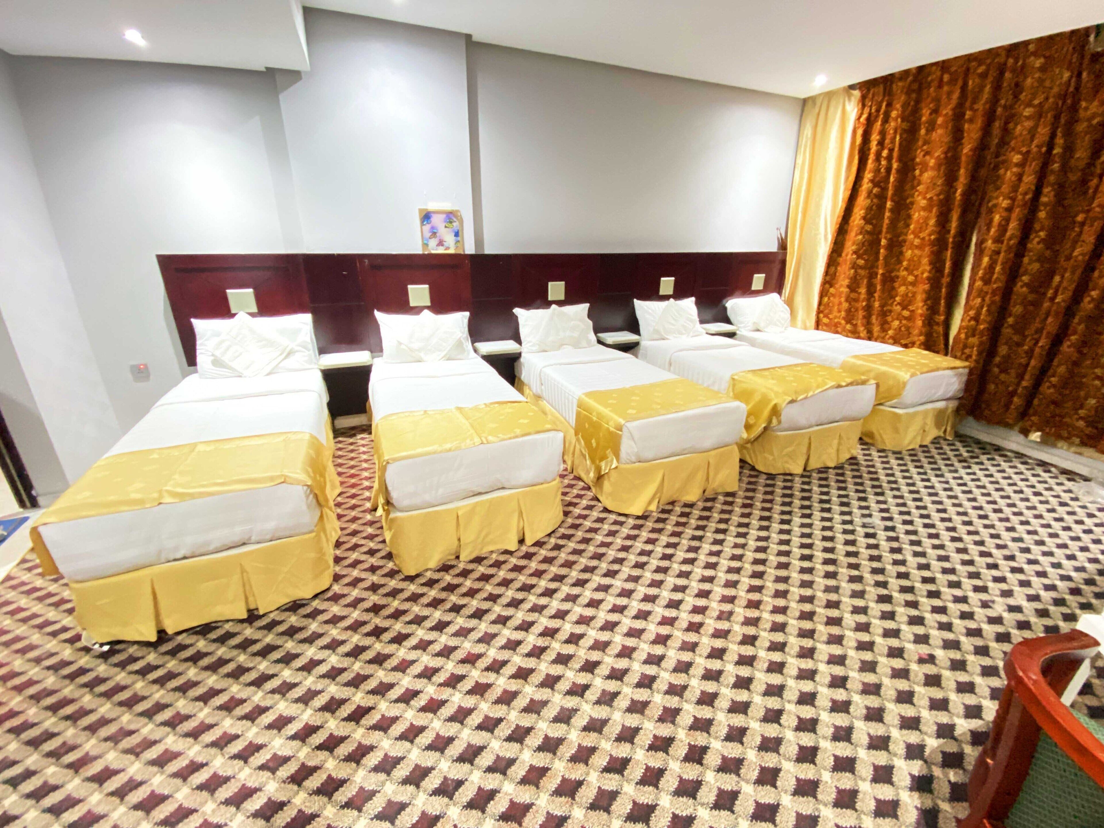 Una habitación de hotel con seis camas dispuestas en dos filas, cada una con un edredón amarillo. La habitación tiene una alfombra a cuadros y un ventanal con cortinas.