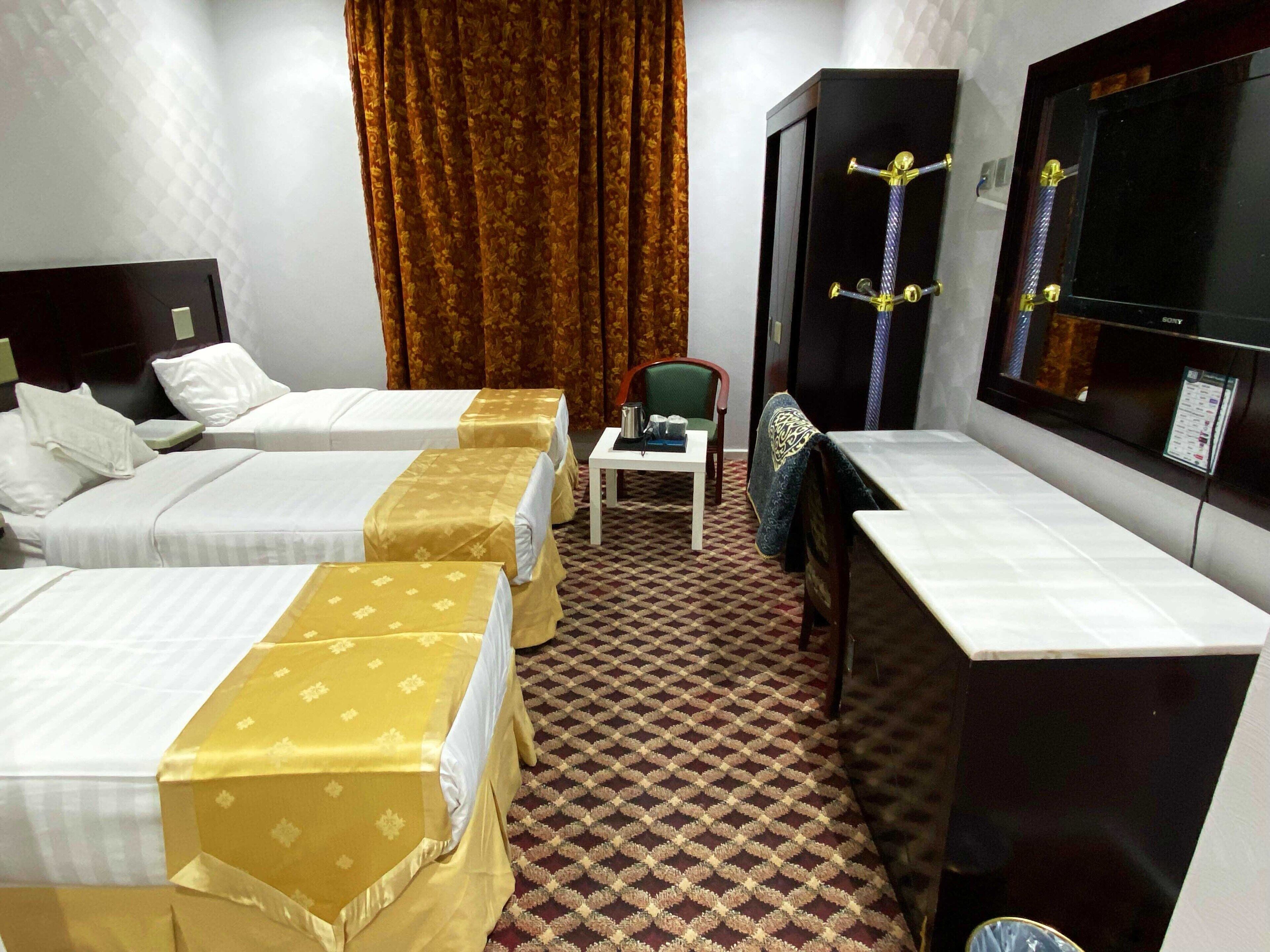 Ein Hotelzimmer mit drei Betten, einem Schreibtisch, einem Sessel, einem Fernseher und einem gemusterten Teppich.