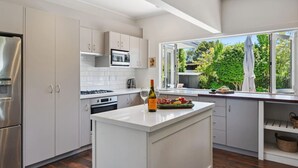 A modern kitchen with white cabinetry, a stainless steel refrigerator, a microwave, and an oven. There's a large window offering a view of greenery outside.