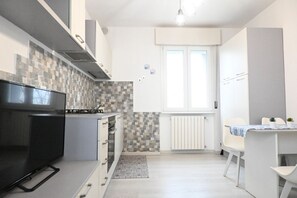 A compact kitchen with a flat-screen TV, a tiled backsplash, white cabinets, and a window with blinds.