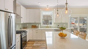 A modern kitchen with white cabinets, stainless steel appliances, a large island with a fruit bowl, and pendant lighting.