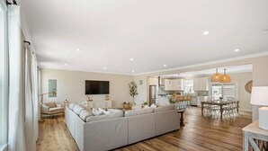 A spacious living room with a large gray sectional sofa, a flat-screen TV, and a dining area with a wooden table and chairs.
