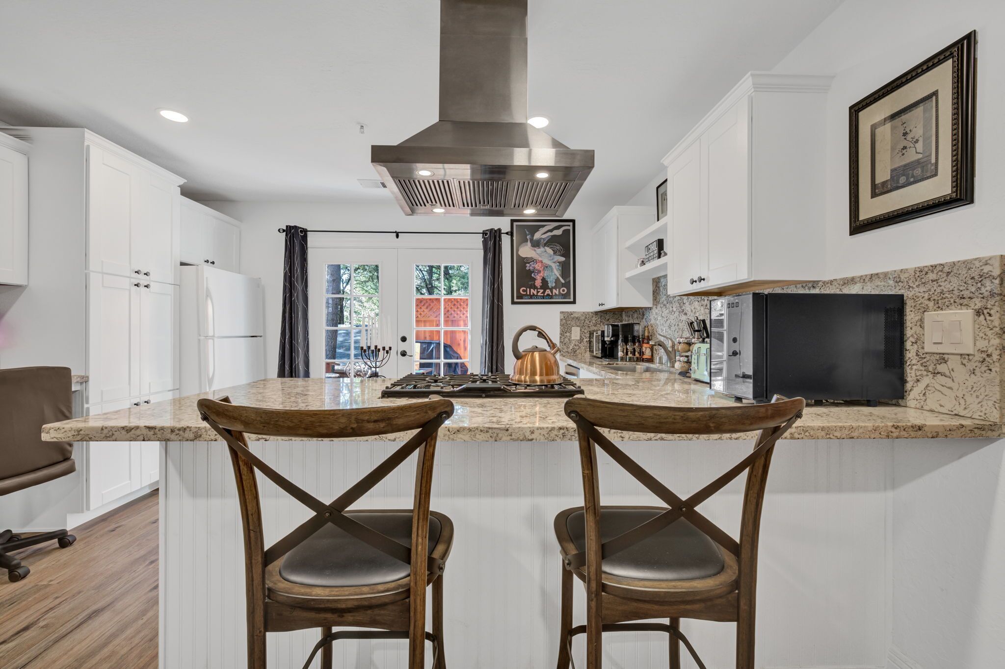 A modern kitchen with white cabinetry, a stainless steel range hood, a copper kettle, and a large island with two wooden chairs.