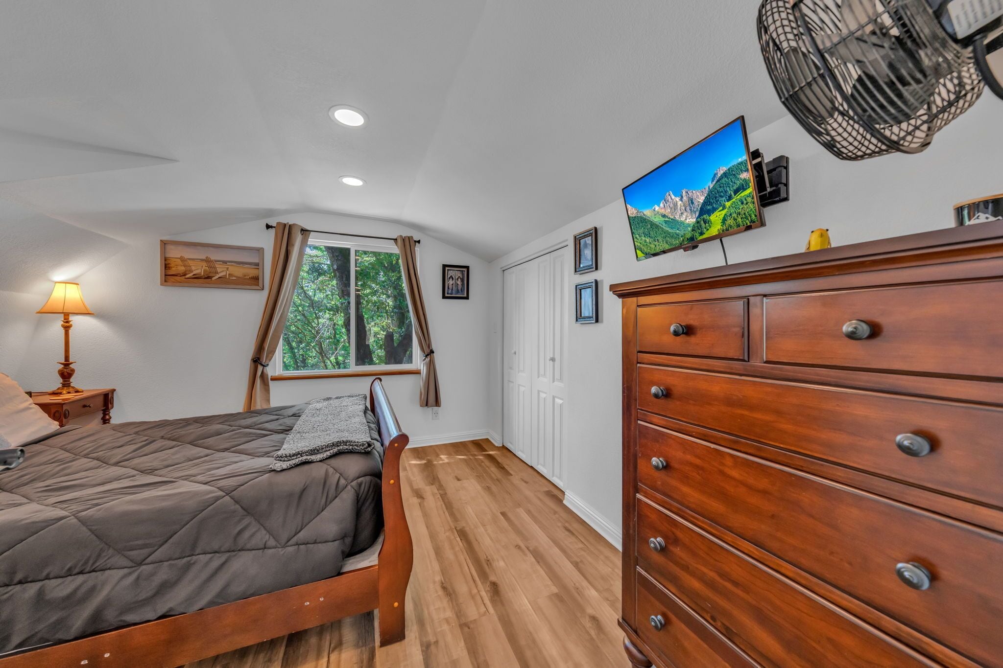 A bedroom with a wooden bed, a dresser, a TV, and a window with curtains.