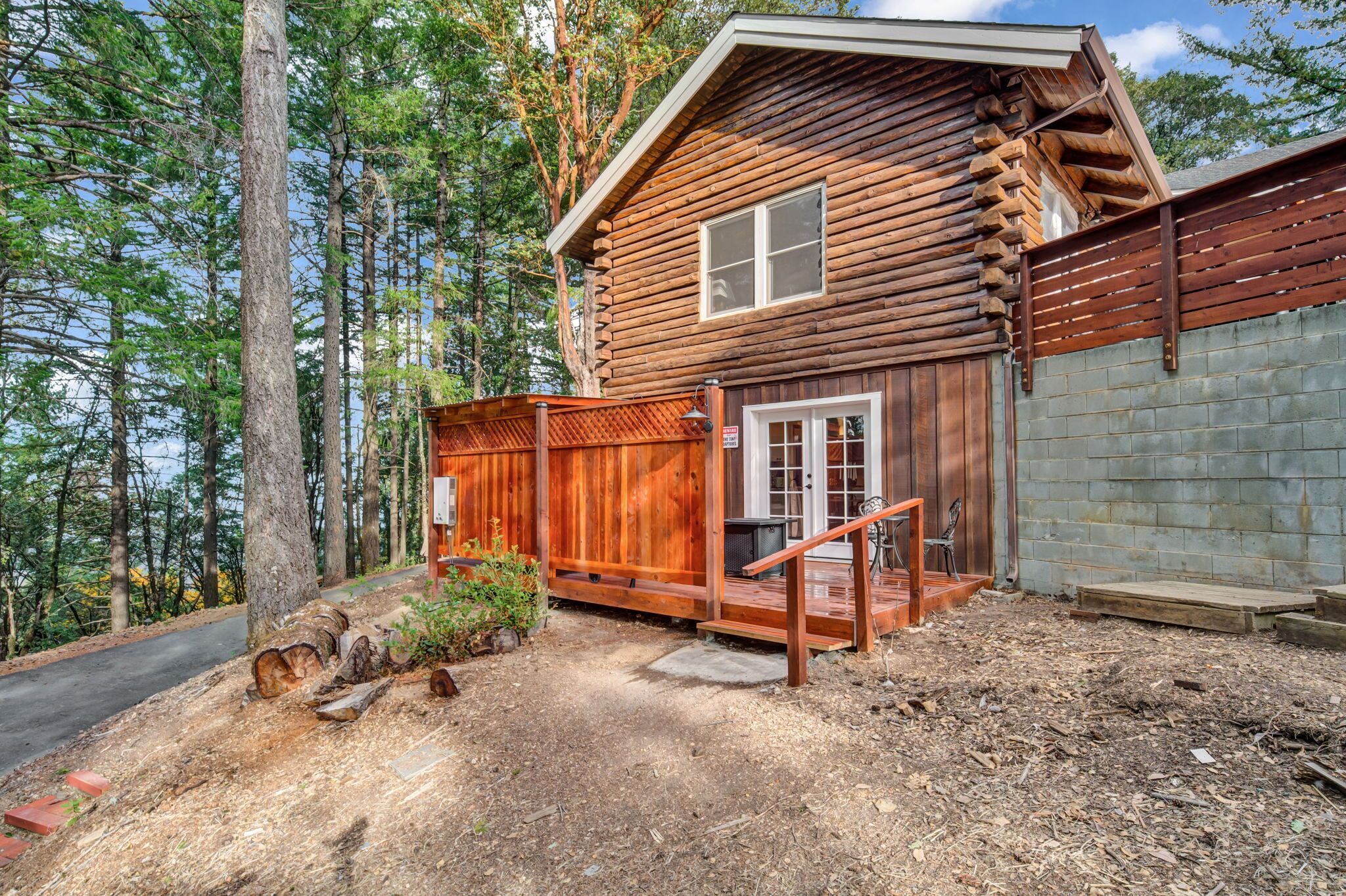 A log cabin with a wooden porch, a brick wall, and a forested backdrop.