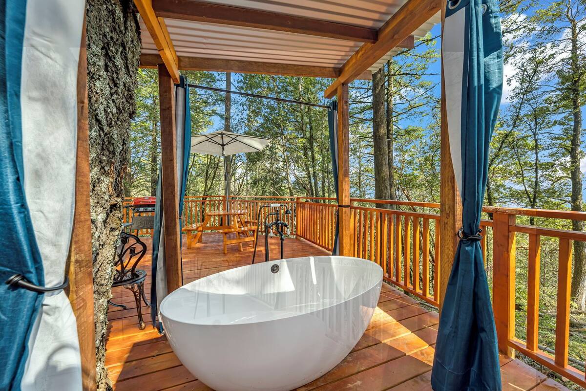 Une terrasse en bois avec une grande baignoire blanche, du mobilier d’extérieur et une vue sur la forêt.