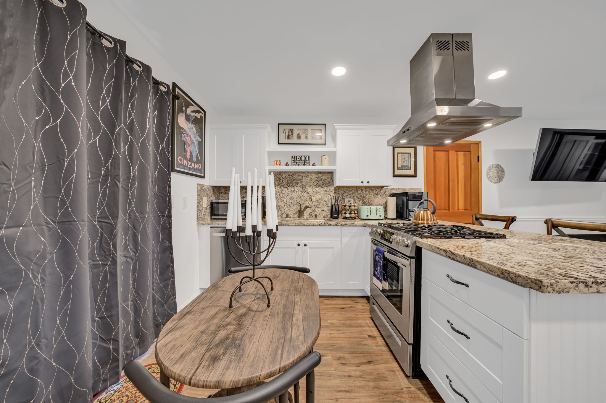 A modern kitchen with white cabinets, a granite countertop, a stainless steel range hood, and a dining area with a wooden table and chairs.