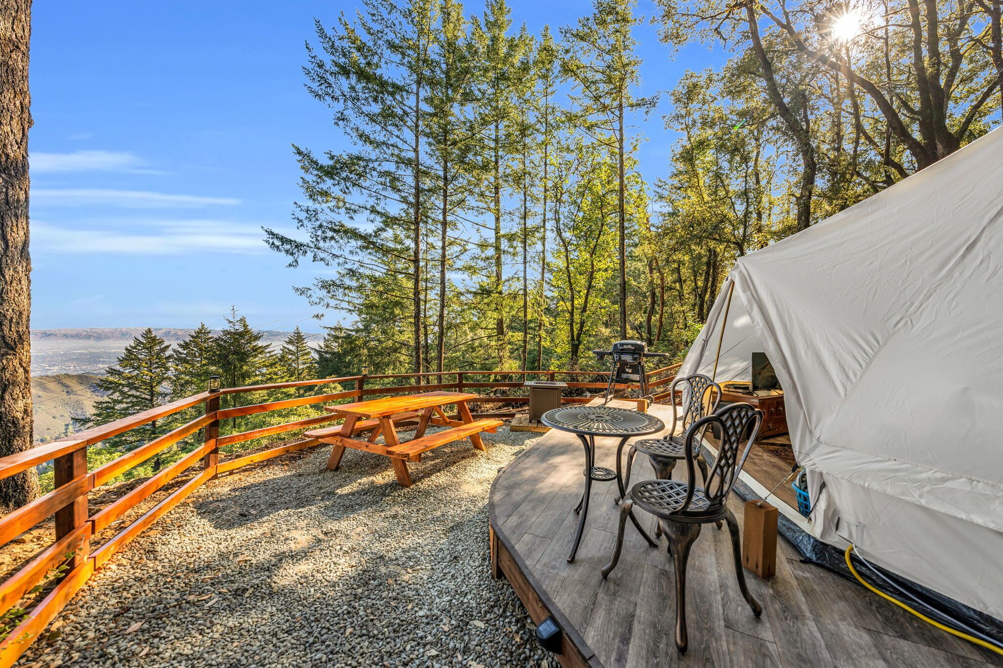 Un espace aménagé avec une tente, une terrasse en bois, une table de pique-nique et des chaises, entouré d’arbres et offrant une vue sur une vallée.