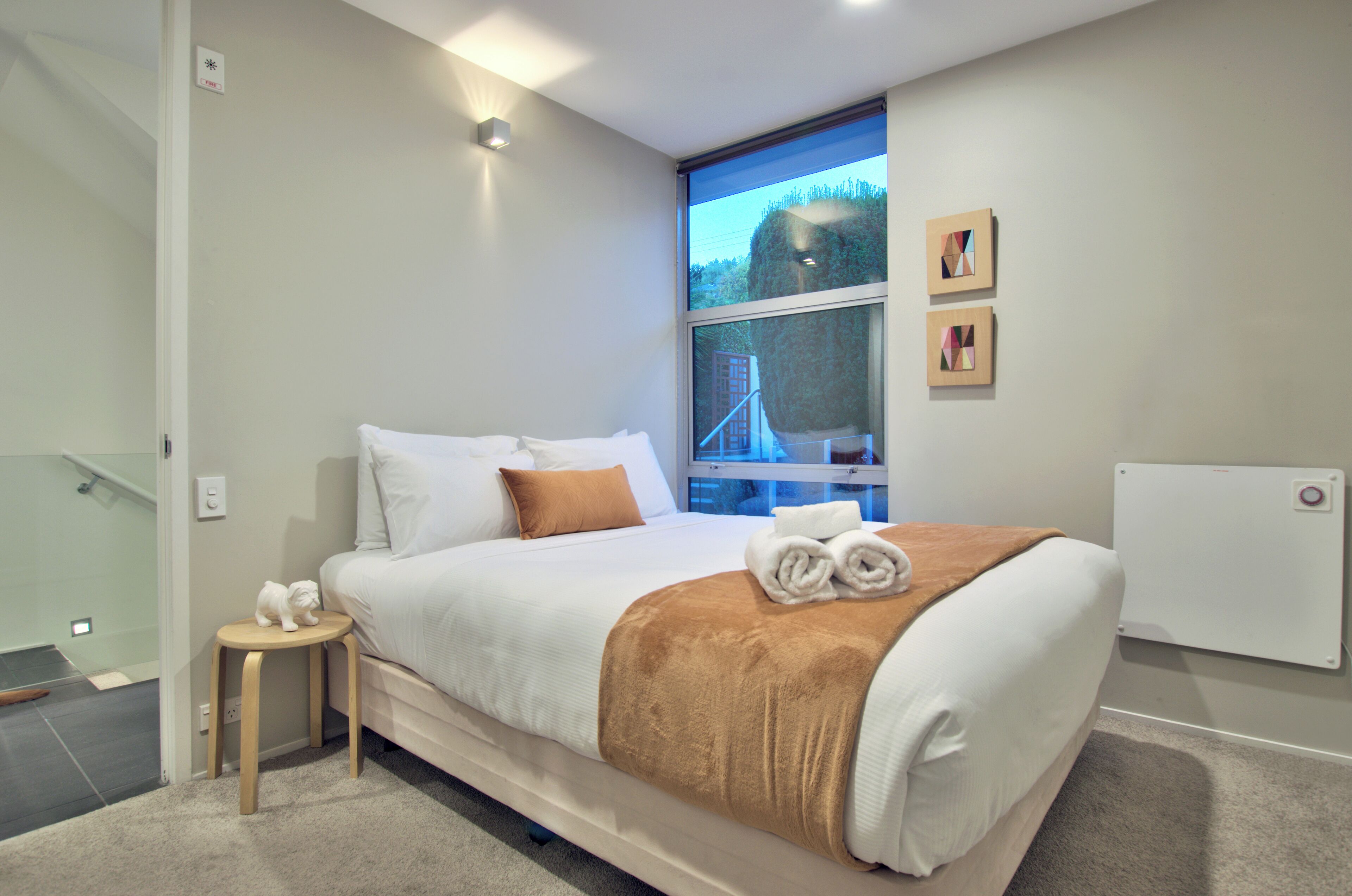 A modern hotel room with a large bed, a small wooden stool, and a view of a bridge through a window.