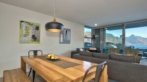Un espace de repas moderne avec une table en bois, des chaises et un saladier rempli de fruits. On y trouve également un canapé gris et, grâce aux grandes fenêtres, une vue sur les montagnes.