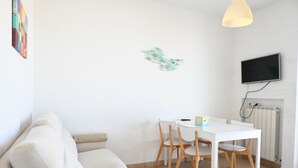 Een witte bank, een eettafel met stoelen, een aan de muur bevestigde televisie en een radiator.