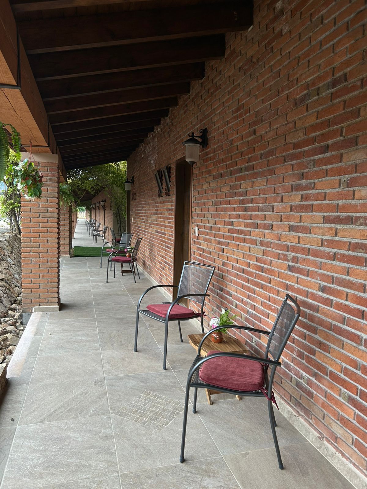 A covered outdoor seating area with brick walls and tiled flooring, featuring metal chairs with cushions.