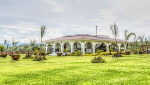 A building with a white facade and a red roof, surrounded by a well-maintained lawn and tropical plants. - Hotel Matea San Isidro  (Tonatico)