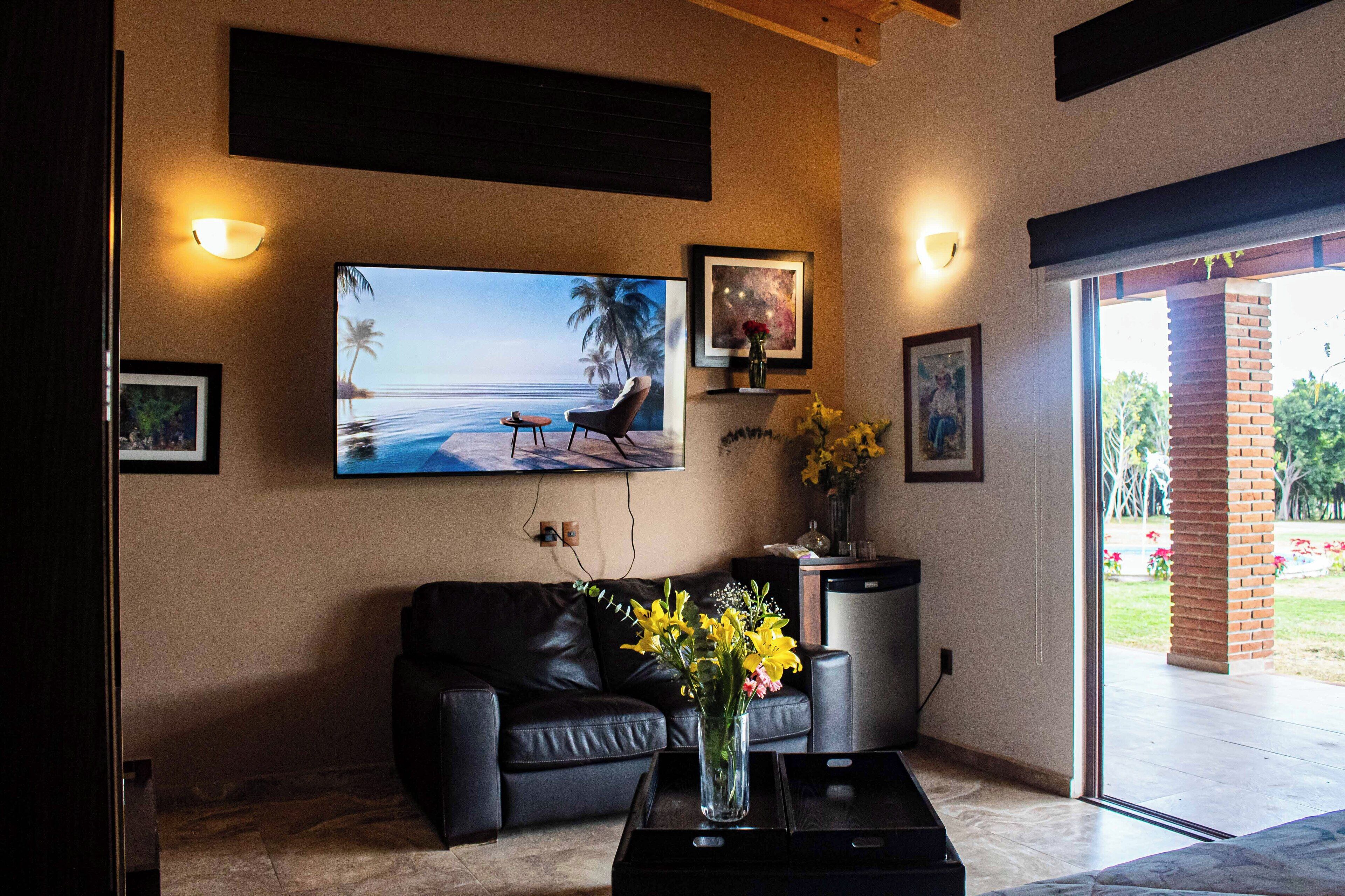 Una sala de estar con un sofá de cuero negro, una mesa de centro con un jarrón de flores, un televisor de pantalla plana montado en la pared y un amplio ventanal con vistas a un jardín.