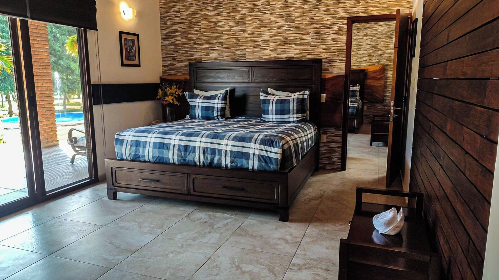 A bedroom with a large bed, a wooden headboard, a small bedside table, and a view of the outdoors through a sliding door.