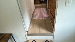 Un pasillo estrecho que conduce a un dormitorio con cama, armario de madera y un pequeño cofre de madera.