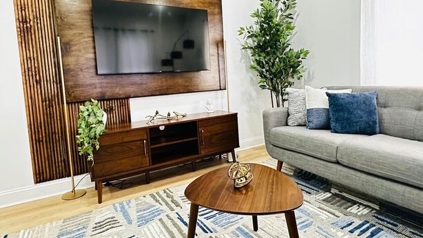 A living room with a flat-screen TV mounted on a wooden panel, a grey sofa with blue cushions, a round wooden coffee table, and a patterned rug. - Feels Like Home - BBQ, Yard, Fire Pit (Fitchburg)