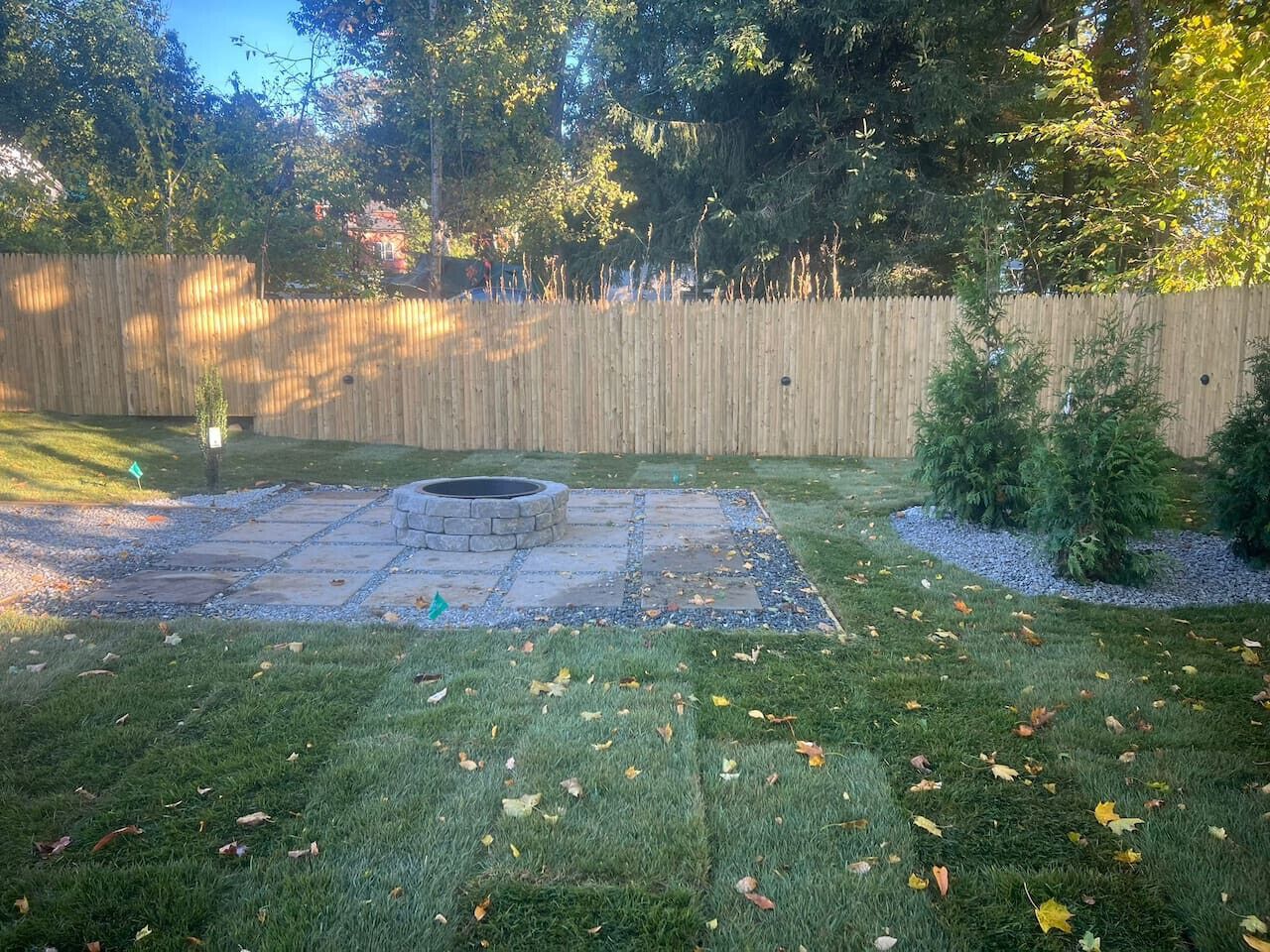 A backyard with a stone fire pit, a wooden fence, and a grassy area with scattered leaves.