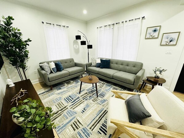 A living room with two grey sofas, a coffee table, a white armchair, a patterned rug, and a floor lamp.