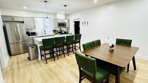 A modern kitchen with a central island, dining area with green upholstered chairs, and a wooden dining table. - Feels Like Home - BBQ, Yard, Fire Pit (Fitchburg)