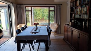A dining area with a table, chairs, a vase with flowers, a bookshelf, and a view through large windows.