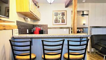 A kitchen with a bar area, three bar stools, a ceiling light fixture, and a framed picture on the wall.