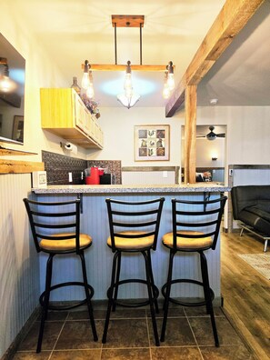 A kitchen with a bar area, three bar stools, a ceiling light fixture, and a framed picture on the wall.
