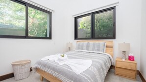 A bedroom with a bed, two windows, a nightstand, a lamp, and a trash bin.