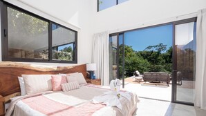 A modern bedroom with a large bed, a wooden headboard, and a view of the outdoors through a sliding glass door.