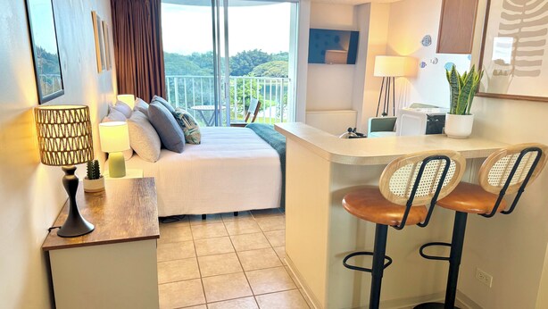 A compact hotel room with a bed, a small desk, two bar stools, a balcony with a view, and a potted plant. - Hilo Studio Retreat with Gorgeous Park View, AC, WiFi, Pool (Hilo)