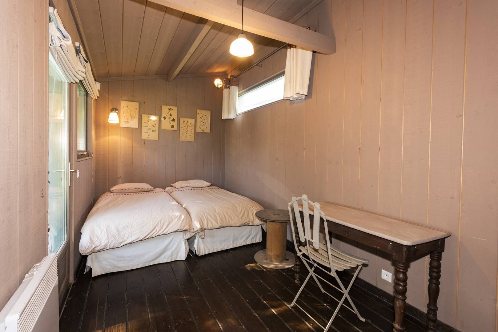 Une petite pièce agréable et chaleureuse, avec deux lits, un bureau en bois et une chaise. On y trouve une fenêtre avec des rideaux, un radiateur et des lampes fixées au mur.