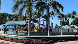 A playground with slides and a covered seating area, surrounded by palm trees and a clear sky.