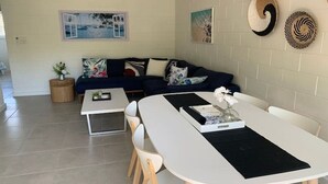 A modern living room with a sofa, a dining table, and chairs. There are wall-mounted decorations and a small side table with a plant.