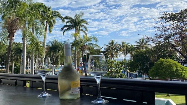 A bar with a bottle and two glasses on a counter, palm trees and a clear sky in the background.