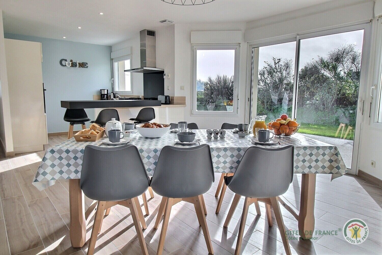 Un coin repas avec une table dressée pour quatre personnes, une cuisine en arrière-plan et une vue sur l’extérieur.