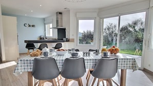 A dining area with a table set for four, a kitchen in the background, and a view of the outdoors. - Ty Roc'h Lanmodez (sleeps 6) (LANMODEZ)
