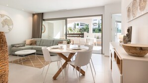 A modern living room with a sofa, dining table, and chairs. There is a large window with a view of greenery outside. - Jardines - Adama 1.3 POOL VIEW (Arona)