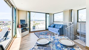Ein modernes Wohnzimmer mit großem Fenster, einem blauen Sofa, einem Esstisch gedeckt mit Tellern und Gläsern, sowie einem Balkon mit Stuhl und Blumentöpfen.