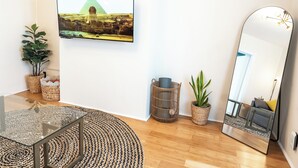 A modern living room with a glass coffee table, a large mirror, a framed picture of a pyramid, and potted plants. - Chic & Comfortable 1 bed apt with Parking. Walking distance JSQ path. Sleeps 4.  (Jersey City)