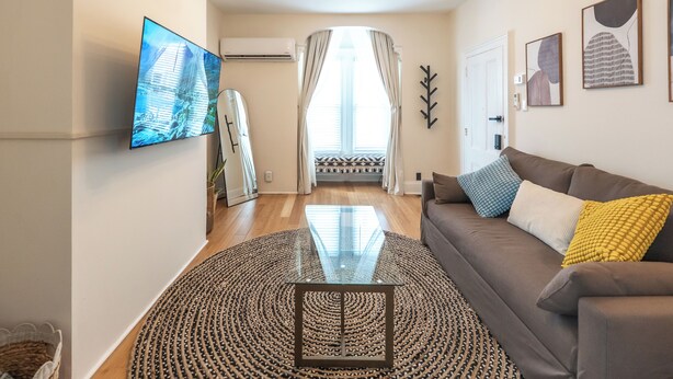 A living room with a sofa, a glass coffee table, a TV, and a large window with curtains. - Chic & Comfortable 1 bed apt with Parking. Walking distance JSQ path. Sleeps 4.  (Jersey City)