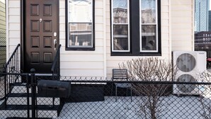 A two-story building with a brown front door, a small porch with a bench, and a visible air conditioning unit. - Chic & Comfortable 1 bed apt with Parking. Walking distance JSQ path. Sleeps 4.  (Jersey City)