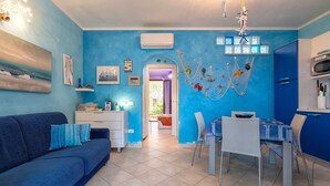 Una sala de estar con un sofá, una mesa de comedor con sillas, una zona de cocina con un refrigerador y una puerta que da a otra habitación.